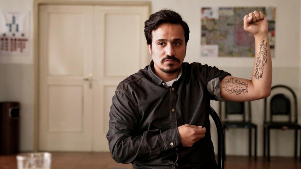 Le point levé, un homme retrousse sa chemise pour montrer les inscriptions tatouées sur son bras. Scène du film "Chroniques de Téhéran".
