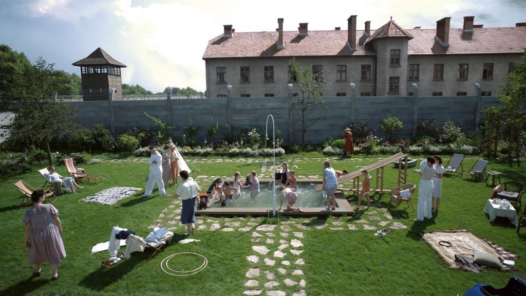 Une ribambelle d'enfants s'amuse dans une piscine au milieu du beau jardin vert. Quelques adultes autour surveillent les jeux des bambins. Un premier plan très joyeux (on peut presque entendre les rires des enfants). Mais derrière le mur de la joyeuse maison se dresse le camp d'Auschwitch. 