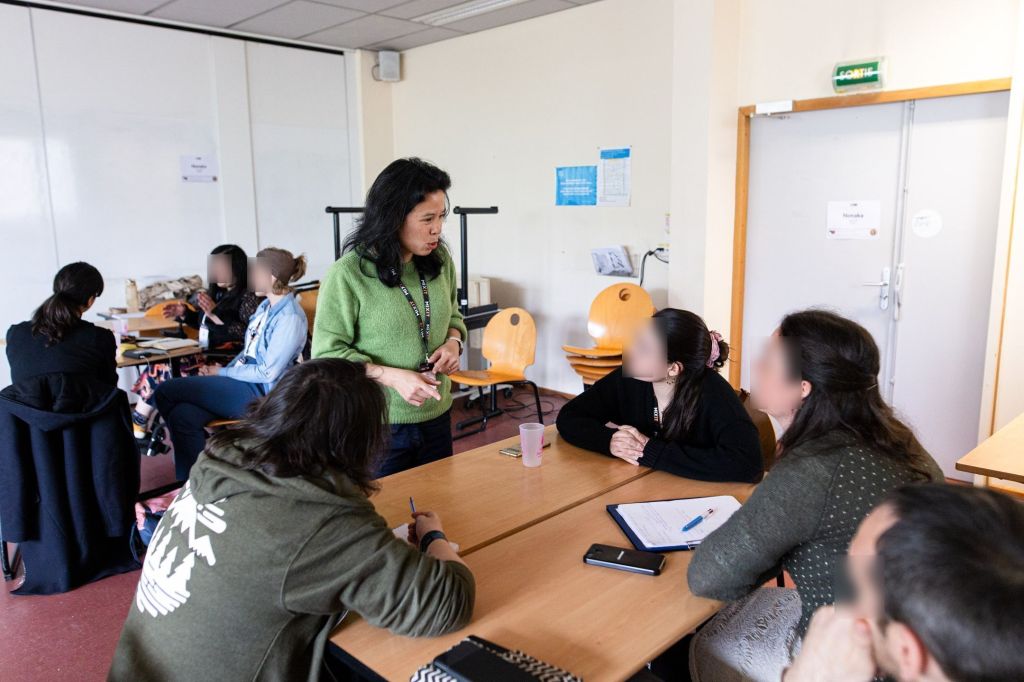 Dans une salle de classe. 
Une personne en pull vert, debout, parle avec un groupe de 4 personnes assise autour d'une table. 
En arrière plan, un autre groupe de trois femmes échangent ensemble.