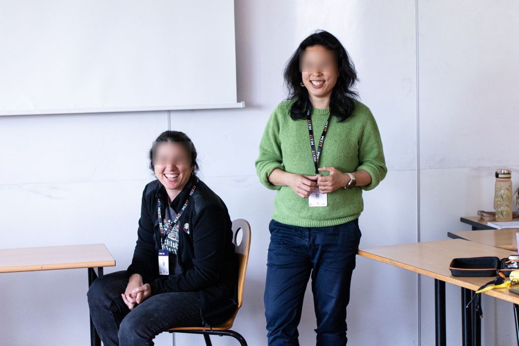 Devant un tableau blanc, une femme est assiste à coté d'une table. Elle regarde le public en riant. Une autre femme est à coté, debout, avec un pull vert et rit aussi dans la même direction.