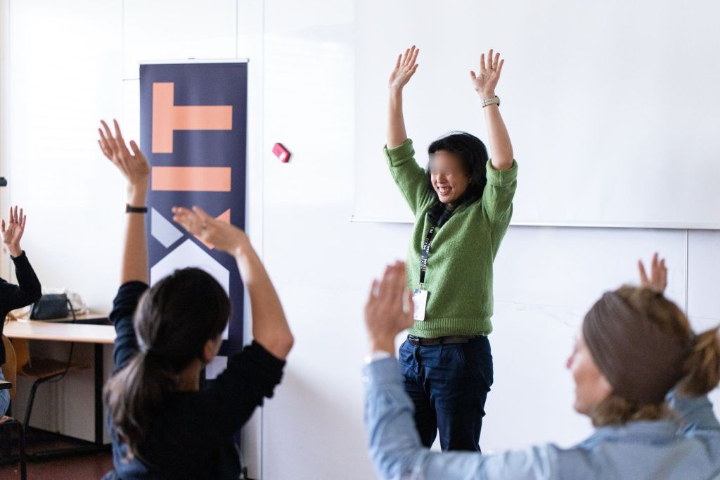 Une personne debout, devant un tableau blanc, lève ses bras en souriant. Trois autres personnes assises ont également les bras levés.